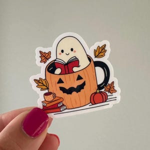 May include: A cute ghost reading a book inside a pumpkin-shaped mug with a black handle. The mug is surrounded by fall leaves and a stack of books. There is a small cup of coffee and a pumpkin next to the mug.