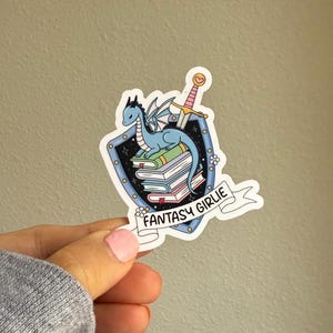 May include: A blue dragon with wings sits on top of a stack of books inside a shield with a pink sword and the text "Fantasy Girlie".