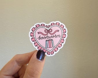 May include: A pink heart-shaped sticker with the word "bookworm" in cursive. The sticker has a pink bow and a stack of books. The sticker is held by a hand.