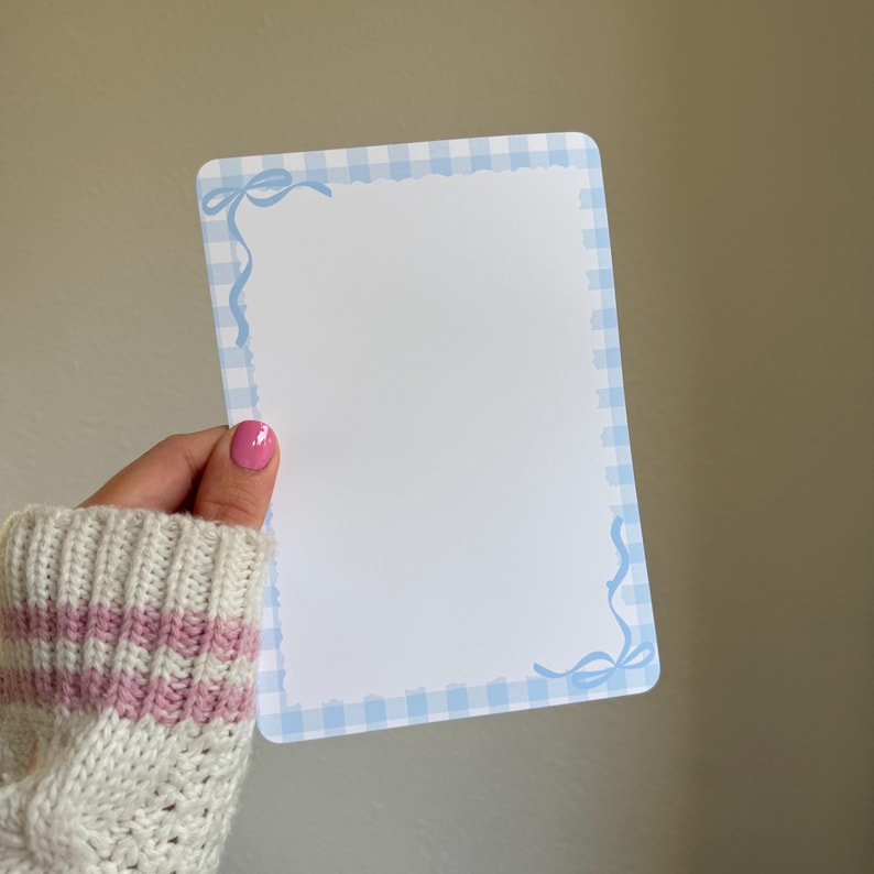 May include: A rectangular white notepad with a light blue gingham border and decorative bows in each corner. The notepad is held by a person wearing a white sweater with pink stripes.