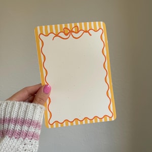 May include: Rectangular notepad with a white interior and a decorative orange border. The notepad has a yellow and white striped background and an orange bow design at the top. The notepad is held by a person wearing a white and pink sweater.