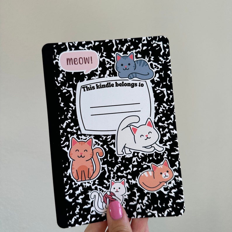 Pu&ograve; includere: Un quaderno a righe bianco e nero con una variet&agrave; di adesivi di gatti colorati. Il quaderno ha il testo "This kindle belongs to" scritto su di esso. Gli adesivi includono un gatto grigio, un gatto bianco, un gatto arancione e un gatto rosa.