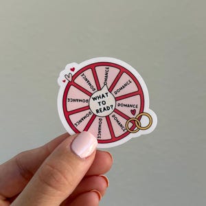 May include: A white sticker with a red and pink wheel design. The wheel is divided into sections with the word "Romance" in each. The center reads "What to Read?" with hearts and gold rings. A hand holds the sticker.