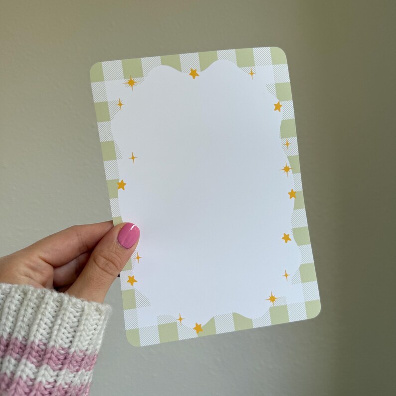 May include: A rectangular notepad with a scalloped edge and a white interior. The border features a light green and white gingham pattern with yellow star accents. The notepad is held by a hand with pink nail polish.