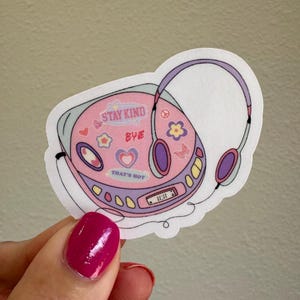 May include: A pink and purple cassette player with headphones attached. The player has a digital display and several buttons. The player has various stickers on it, including "Stay Kind", "Bye", and "That's Hot".