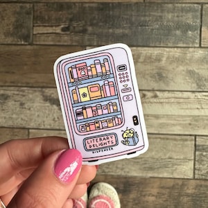 May include: A pink and blue sticker of a vending machine filled with books. The vending machine has a sign that says "Literary Delights Dispenser". A small bee with a book is on the bottom right corner of the sticker.