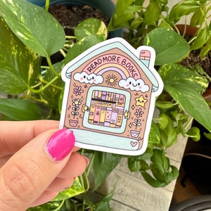 May include: A colorful sticker in the shape of a house with the text "READ MORE BOOKS" above a rainbow. The house features a window filled with books, flowers, and a smiling sun. The sticker is held in front of a background of green leaves.