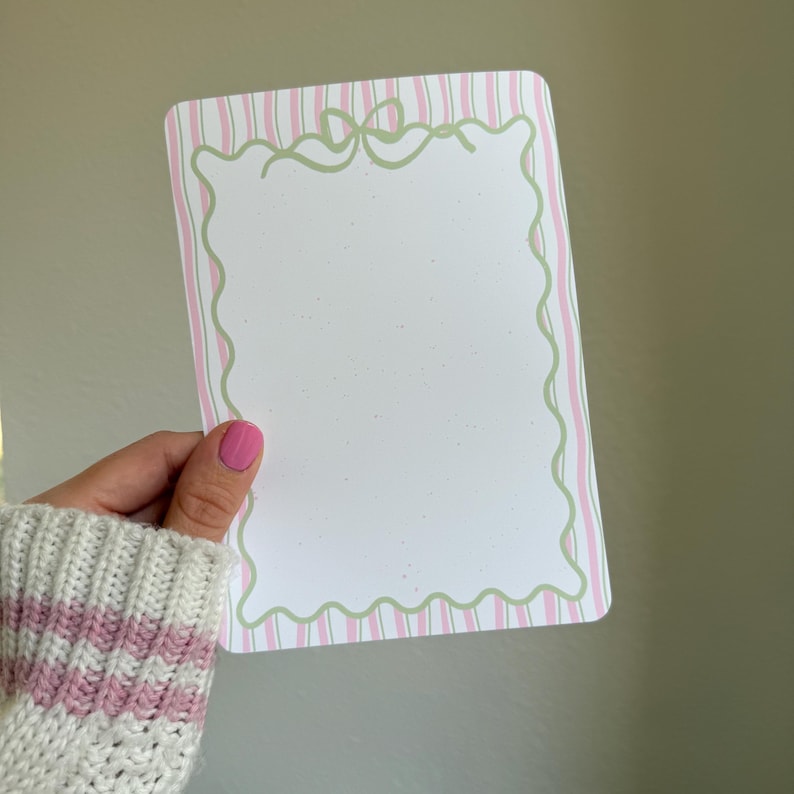 May include: A rectangular notepad with a white interior and a pink and green striped border. The border features a wavy green line and a bow design at the top. The notepad is held by a person wearing a white sweater with pink stripes.