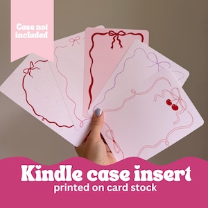 May include: A hand holds several pink Kindle case inserts with red ribbon and cherry designs. The text "Kindle case insert printed on card stock" is at the bottom, and "Case not included" is at the top.