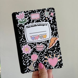 Pu&ograve; includere: Un quaderno nero e bianco con un motivo a scacchi nero e bianco. Il quaderno ha diversi adesivi colorati, tra cui cuori, un fulmine, un fiore, una faccina sorridente, una bocca e una stella. Il quaderno ha un'etichetta che dice "This kindle belongs to" con una riga vuota per scrivere un nome.