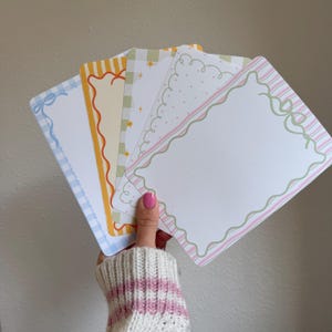 May include: A hand holds a fan of decorative rectangular cards. Each card features a different border design, including gingham, stripes, and wavy lines, in pastel colors like blue, yellow, green, and pink. The cards have a white interior.
