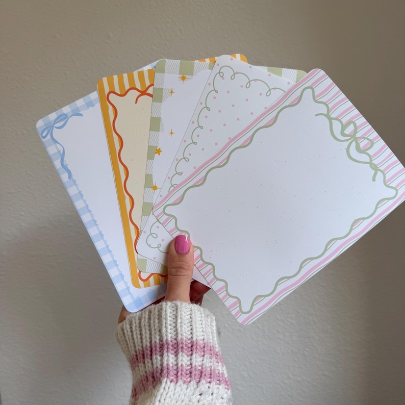 May include: A hand holds a fan of decorative rectangular cards. Each card features a different border design, including gingham, stripes, and wavy lines, in pastel colors like blue, yellow, green, and pink. The cards have a white interior.