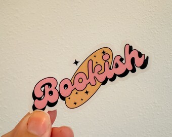 May include: A clear sticker with the word "Bookish" in a retro font, outlined in black and pink, with a light orange background and star accents. The sticker is held by a hand.