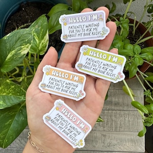 May include: Three colorful stickers with the text "Hello I'm" and "Patiently waiting for you to ask me about my book recs". The stickers are decorated with flowers and books.
