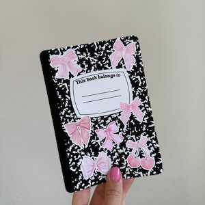 Pu&ograve; includere: Un quaderno nero e bianco con un adesivo a forma di fiocco rosa e bianco e il testo "Questo libro appartiene a" sulla copertina.