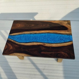May include: A dark wood coffee table with a blue resin river running through the center. The table has a natural wood finish and gold metal legs.