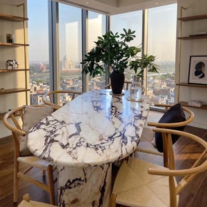 Peut inclure: Une table à manger ovale avec un plateau en marbre veiné blanc et violet et une base cylindrique assortie. Quatre chaises en bois clair avec des assises tressées entourent la table. Un vase noir avec du feuillage vert et une carafe d'eau.