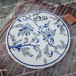 May include: Round white marble table top with a blue and green floral design inlaid with mother of pearl. The design features intricate floral patterns with blue outlines and green and blue mother of pearl accents.