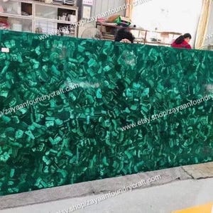 Puede incluir: Gran losa rectangular de piedra de malaquita, que muestra un color verde vibrante con patrones intrincados. La superficie pulida refleja la luz, resaltando las variaciones naturales de la piedra. Adecuada para encimeras u otras aplicaciones de diseño.