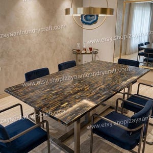 Op de afbeelding: Een rechthoekige eettafel met een gepolijst tijgeroog stenen oppervlak en een metalen onderstel, omringd door zes marineblauwe gestoffeerde stoelen met metalen frames. Een ronde gouden hanglamp met een blauw agaat middenstuk hangt erboven.