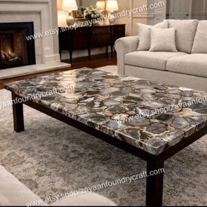 May include: A rectangular coffee table with a dark brown wooden frame and a top made of polished, irregularly shaped, gray and brown stones. The table is in a living room with a fireplace and neutral-toned furniture.