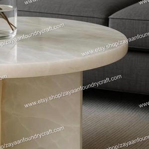 May include: A modern, round coffee table with a light-colored, marble-like top and a rectangular base. The table is set against a gray sofa and a neutral-toned rug. The website address www.etsy.shop/zayaanfoundrycraft.com is visible.