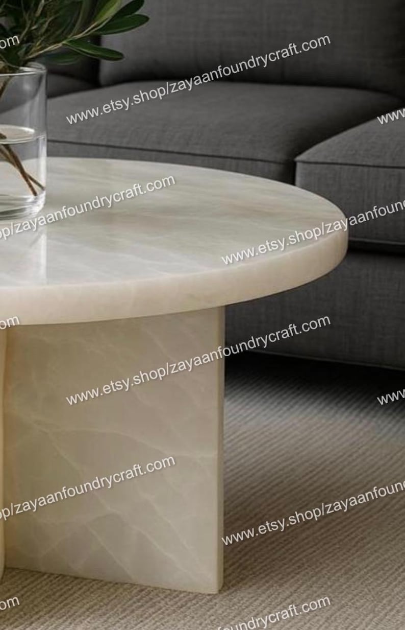 May include: A modern, round coffee table with a light-colored, marble-like top and a rectangular base. The table is set against a gray sofa and a neutral-toned rug. The website address www.etsy.shop/zayaanfoundrycraft.com is visible.