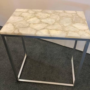 May include: A white quartz stone top end table with a silver metal frame. The table has a modern design and would be a great addition to any home.