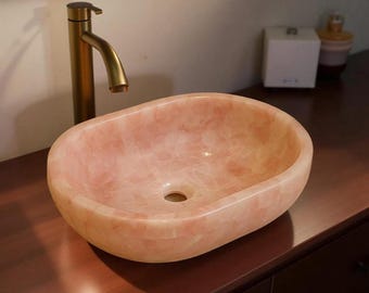 May include: Oval-shaped pink stone bathroom sink with a gold faucet. The sink has a smooth, polished surface and a drain hole. The faucet is a modern design with a curved spout. The sink is set on a dark brown wooden surface.