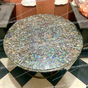 Könnte beinhalten: Ein runder Couchtisch mit Mosaikplatte. Die Tischplatte ist mit schillernden Teilen bedeckt, wodurch ein schimmernder Effekt entsteht. Der Tisch hat ein bronzefarbenes Metallgestell. Der Durchmesser des Tisches beträgt ca. 122 cm.