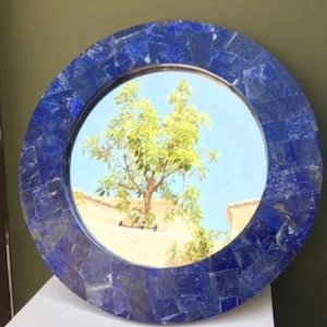 Op de afbeelding: Een ronde spiegel met een lijst gemaakt van blauwe lapis lazuli stenen tegels. De spiegel reflecteert een boom en een gebouw.