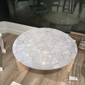 Può includere: Tavolino rotondo con piano bianco a mosaico. Il piano è composto da pezzi di forma irregolare, creando un motivo unico. La base è in legno chiaro, che offre un contrasto naturale.
