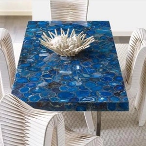 May include: A blue agate dining table with a white centerpiece and four white chairs. The table top is made of polished agate stones, creating a unique and eye-catching design.