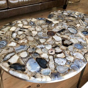 May include: Two round agate coffee tables with a silver metal base. The tabletops are made of natural agate stone with a variety of brown, gray, and white colors.