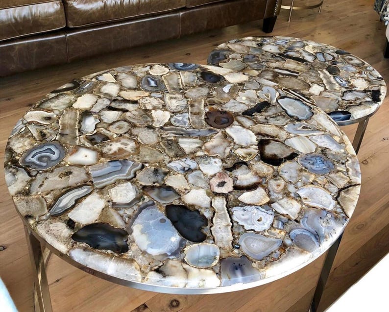 May include: Two round agate coffee tables with a silver metal base. The tabletops are made of natural agate stone with a variety of brown, gray, and white colors.