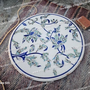 May include: Round white marble table top with a floral design inlaid with blue and green mother of pearl. The table top is approximately 24 inches in diameter.