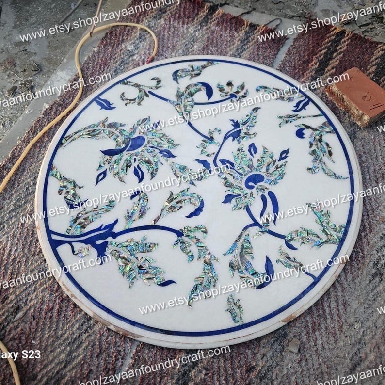 May include: Round white marble table top with a floral design inlaid with blue and green mother of pearl. The table top is approximately 24 inches in diameter.