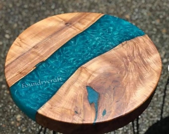 May include: Round wooden table top with a turquoise blue epoxy resin river running through the centre. The resin has a sparkly, glittery effect. The text "Foundrycraft" is visible in the resin.