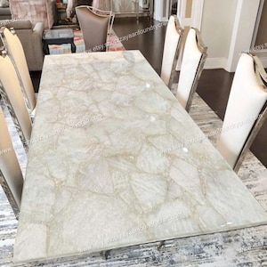 May include: A white quartz dining table with a unique, natural stone pattern. The table is surrounded by six white chairs with silver legs. The table is approximately 7 feet long and 3 feet wide.