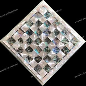 May include: A white and black checkerboard made of mother of pearl inlay. The board is surrounded by a white border with a thin black line.