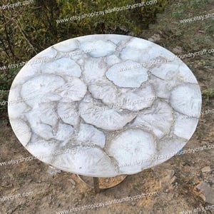 May include: A round white table with a unique agate-like design. The table top is made of white agate and has a natural, organic look. The table is supported by a silver metal base.