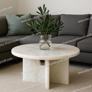 May include: A round, off-white marble coffee table with a unique pedestal base. A clear glass vase with green leafy branches sits on the table. A gray sofa and a green pillow are in the background.