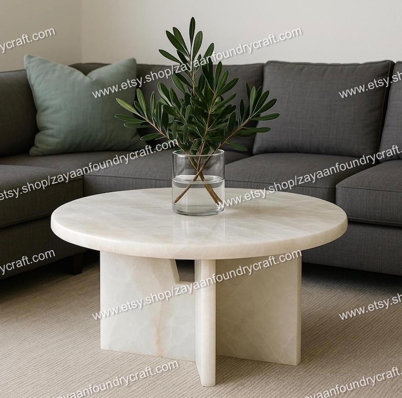 May include: A round, off-white marble coffee table with a unique pedestal base. A clear glass vase with green leafy branches sits on the table. A gray sofa and a green pillow are in the background.