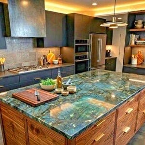 May include: A kitchen island with a polished blue and green labradorite stone countertop and walnut wood cabinetry. The island features a wooden cutting board, a glass oil bottle, and small ceramic bowls. Dark cabinets and stainless steel appliances.