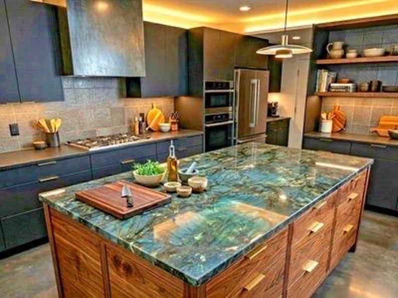 May include: A kitchen island with a polished blue and green labradorite stone countertop and walnut wood cabinetry. The island features a wooden cutting board, a glass oil bottle, and small ceramic bowls. Dark cabinets and stainless steel appliances.