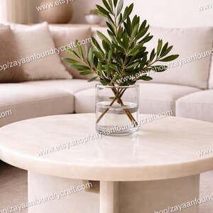 May include: A round, cream-colored marble coffee table with a cylindrical base. A clear glass vase with green leafy branches sits on top. The image also shows a beige sofa and a lamp.