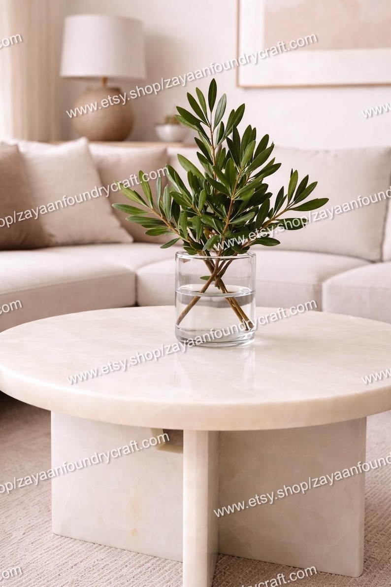 May include: A round, cream-colored marble coffee table with a cylindrical base. A clear glass vase with green leafy branches sits on top. The image also shows a beige sofa and a lamp.