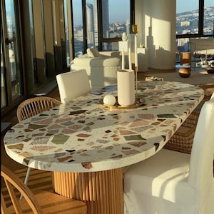 May include: A white oval table with a terrazzo top and a wooden base. The table is surrounded by white chairs and a wicker chair. The table is set with a white vase and a gold tray with a white decorative object.