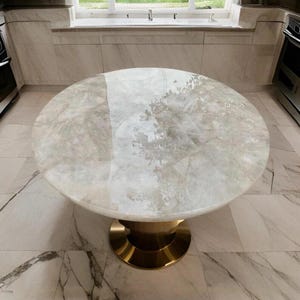 May include: A round dining table with a light-colored, patterned top and a gold-colored pedestal base. The table is in a kitchen with marble flooring and a window in the background. The table is approximately 48 inches in diameter.