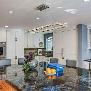 May include: A rectangular crystal chandelier hangs above a kitchen island with a dark blue and black granite countertop. On the island sits a silver textured vase with purple flowers, a blue rectangular lidded dish, and two yellow oranges.